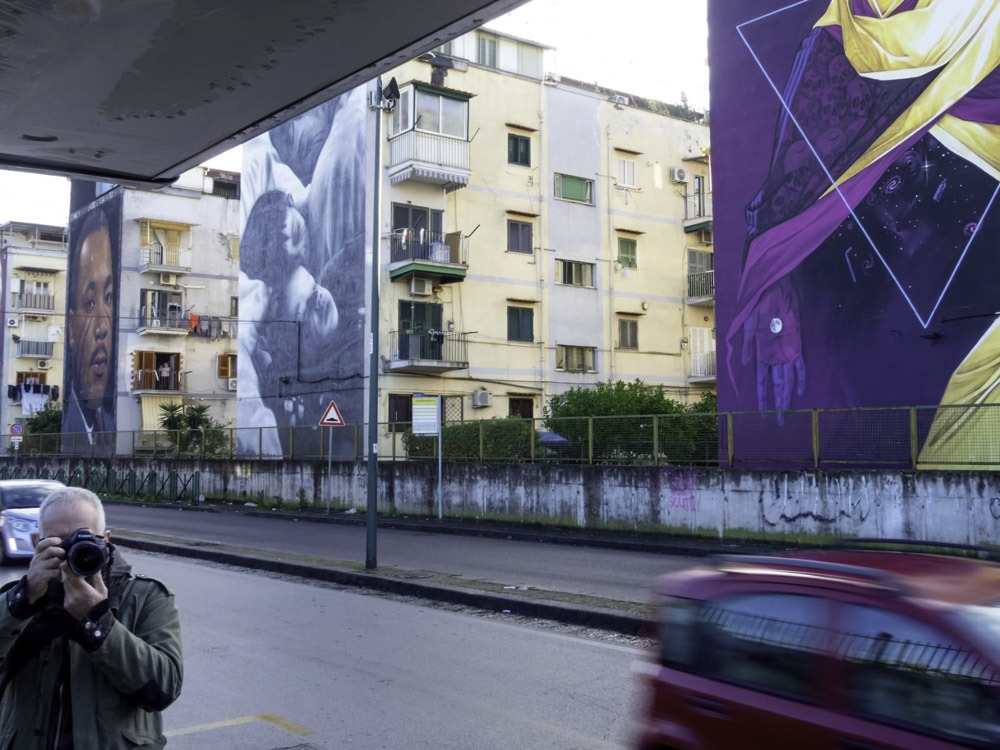 La street art può trasformare tutto ciò che è per strada in un’opera inedita. E lo sa bene Jorit che, con i suoi murales, da 20 anni è entrato a far parte del tessuto urbano di Napoli cambiando radicalmente il volto ai quartieri, dando così un significato diverso allo spazio abitato. Proprio per il suo potere trasformativo, la street art di Jorit si contraddistingue per una forte componente sociale, indissolubile dal contesto di origine. Nel rapporto tra lui e la città c’è una profonda identificazione ed empatia. Come un “Caravaggio contemporaneo” Jorit ha scelto anche gente comune per le sue opere: da non perdere assolutamente a Ponticelli il volto della bambina rom, o quello di San Gennaro all’ingresso di Forcella, a cui Jorit ha dato il volto di un suo amico carrozziere. Dal centro storico fino all’estrema periferia suburbana: da Piazzetta Eduardo De Filippo, con il bellissimo trittico sulla facciata del Teatro san Ferdinando, fino al volto gigantesco di un’altra icona della cultura popolare partenopea: Maradona a Quarto. MA CHI È JORIT? Il nome d’arte Jorit Agoch è composto dall’unione del suo vero nome di battesimo e la sua tag. All’anagrafe Jorit Ciro Cerullo, classe ’90, con padre partenopeo e madre olandese, nato e cresciuto nella periferia Nord di Napoli, a Quarto. La svolta nella sua poetica è senza dubbio il suo viaggio in Africa nel 2005: scopre la Tanzania, i suoi paesaggi, il suo popolo e ne rimane affascinato, tanto da tornarci più volte, anche per studiare per la scuola d’arte Tinga Tinga di Dar es Salaam. L’Africa lo segnerà a vita e il suo stile subisce un cambiamento radicale, passando da rappresentazioni semplici e writing allo studio del volto umano, della sua rappresentazione e delle emozioni che esso può trasmettere. I volti diventano il soggetto principale della sua ricerca: c’è qualcosa di profondo nel nostro essere umani. Rappresentare un volto non è semplicemente dipingere una persona, ma mettere l’osservatore a tu per tu con se stesso: come possiamo provare odio quando guardiamo una persona negli occhi? Tutti i volti dipinti da Jorit hanno una caratteristica: due strisce rosse sulle guance. Durante i suoi viaggi in Africa l’artista entra in contatto con persone appartenenti a diverse tribù locali ed approfondisce il rito della scarificazione. Alcune tribù in Tanzania e Kenya segnano i volti degli appartenenti alla tribù stessa come simbolo di fratellanza ed accettazione oppure come simbolo di ingresso nell’età adulta; la scarificazione consiste in diverse tecniche, ma la più usata è costituita da tagli di varia forma e lunghezza: ogni tribù ha il proprio rito e questo permette anche di riconoscere la tribù di appartenenza di una persona. Affascinato da questo concetto, Jorit ha deciso di trasportarlo all’interno dei suoi muri creando quella che lui chiama Tribù Umana o Human Tribe: il colore della pelle, degli occhi, dei capelli, la tua età, il fatto di essere uomo o donna non ti rendono migliore o peggiore, siamo tutti esseri umani ed apparteniamo ad una grande tribù. I segni rossi inconfondibili sui volti che Jorit imprime quasi “a ferro e fuoco”, sorta di meta-tatuaggio, simboleggiano quindi l’unità della tribù opposta alla singolarità dell’individuo e sono presenti in tutti i volti/murales dell’artista. Così da Forcella al Vomero, da Poggioreale a San Giovanni a Teduccio, passando pe il mare di Bacoli fino al quartiere dell’Avvocata e al Cardarelli, possiamo sentirci anche noi parte di questa grande Human Tribe. IL MESSAGGIO SEGRETO SUI SUOI MURALES. Nei i murales di Jorit sono nascosti dei messaggi, parole e frasi che ne ampliano il significato: nomi di persone del luogo, testimonianze e motti rivoluzionari celati sapientemente dall’artista. Alcuni tra i messaggi nascosti più significativi sono: «Hasta siempre» nel murale di Diego Armando Maradona, «Tagliateci la testa col machete» e «Meglio sparare che sparire» nel murale di Ernesto Che Guevara, «Le vele urlano!» e «Scampia chiede lavoro» nel murale di Pier Paolo Pasolini e «Socialismo o barbarie?» in quello di Salvador Allende. I messaggi sono nascosti ovunque sul volto ma quelli più importanti si trovano negli occhi, dove le persone inizialmente indirizzano il loro sguardo, creando empatia con il soggetto.
