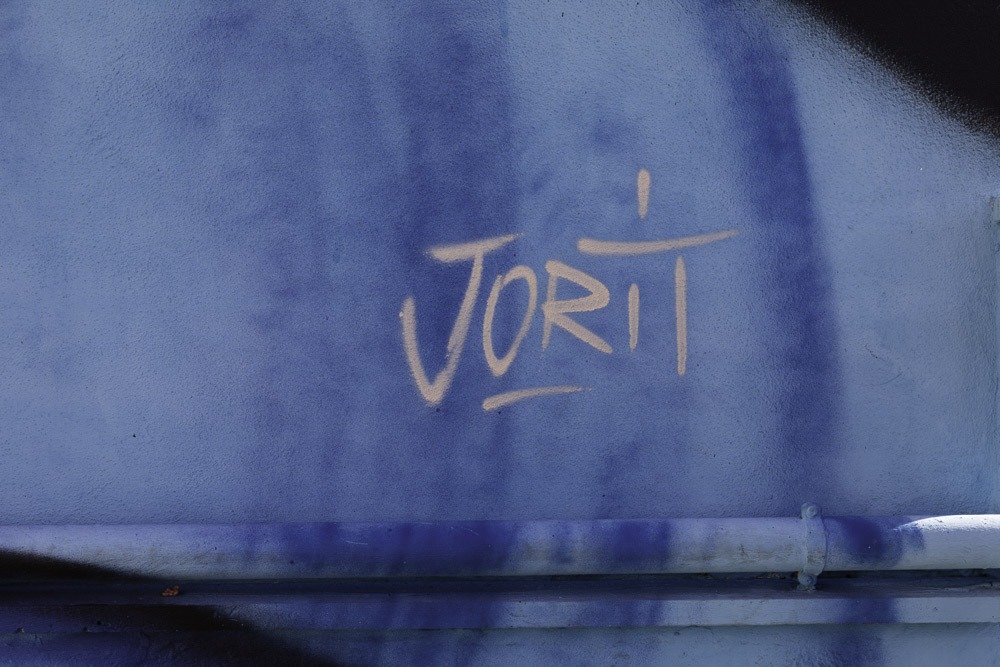 La street art può trasformare tutto ciò che è per strada in un’opera inedita. E lo sa bene Jorit che, con i suoi murales, da 20 anni è entrato a far parte del tessuto urbano di Napoli cambiando radicalmente il volto ai quartieri, dando così un significato diverso allo spazio abitato. Proprio per il suo potere trasformativo, la street art di Jorit si contraddistingue per una forte componente sociale, indissolubile dal contesto di origine. Nel rapporto tra lui e la città c’è una profonda identificazione ed empatia. Come un “Caravaggio contemporaneo” Jorit ha scelto anche gente comune per le sue opere: da non perdere assolutamente a Ponticelli il volto della bambina rom, o quello di San Gennaro all’ingresso di Forcella, a cui Jorit ha dato il volto di un suo amico carrozziere. Dal centro storico fino all’estrema periferia suburbana: da Piazzetta Eduardo De Filippo, con il bellissimo trittico sulla facciata del Teatro san Ferdinando, fino al volto gigantesco di un’altra icona della cultura popolare partenopea: Maradona a Quarto. MA CHI È JORIT? Il nome d’arte Jorit Agoch è composto dall’unione del suo vero nome di battesimo e la sua tag. All’anagrafe Jorit Ciro Cerullo, classe ’90, con padre partenopeo e madre olandese, nato e cresciuto nella periferia Nord di Napoli, a Quarto. La svolta nella sua poetica è senza dubbio il suo viaggio in Africa nel 2005: scopre la Tanzania, i suoi paesaggi, il suo popolo e ne rimane affascinato, tanto da tornarci più volte, anche per studiare per la scuola d’arte Tinga Tinga di Dar es Salaam. L’Africa lo segnerà a vita e il suo stile subisce un cambiamento radicale, passando da rappresentazioni semplici e writing allo studio del volto umano, della sua rappresentazione e delle emozioni che esso può trasmettere. I volti diventano il soggetto principale della sua ricerca: c’è qualcosa di profondo nel nostro essere umani. Rappresentare un volto non è semplicemente dipingere una persona, ma mettere l’osservatore a tu per tu con se stesso: come possiamo provare odio quando guardiamo una persona negli occhi? Tutti i volti dipinti da Jorit hanno una caratteristica: due strisce rosse sulle guance. Durante i suoi viaggi in Africa l’artista entra in contatto con persone appartenenti a diverse tribù locali ed approfondisce il rito della scarificazione. Alcune tribù in Tanzania e Kenya segnano i volti degli appartenenti alla tribù stessa come simbolo di fratellanza ed accettazione oppure come simbolo di ingresso nell’età adulta; la scarificazione consiste in diverse tecniche, ma la più usata è costituita da tagli di varia forma e lunghezza: ogni tribù ha il proprio rito e questo permette anche di riconoscere la tribù di appartenenza di una persona. Affascinato da questo concetto, Jorit ha deciso di trasportarlo all’interno dei suoi muri creando quella che lui chiama Tribù Umana o Human Tribe: il colore della pelle, degli occhi, dei capelli, la tua età, il fatto di essere uomo o donna non ti rendono migliore o peggiore, siamo tutti esseri umani ed apparteniamo ad una grande tribù. I segni rossi inconfondibili sui volti che Jorit imprime quasi “a ferro e fuoco”, sorta di meta-tatuaggio, simboleggiano quindi l’unità della tribù opposta alla singolarità dell’individuo e sono presenti in tutti i volti/murales dell’artista. Così da Forcella al Vomero, da Poggioreale a San Giovanni a Teduccio, passando pe il mare di Bacoli fino al quartiere dell’Avvocata e al Cardarelli, possiamo sentirci anche noi parte di questa grande Human Tribe. IL MESSAGGIO SEGRETO SUI SUOI MURALES. Nei i murales di Jorit sono nascosti dei messaggi, parole e frasi che ne ampliano il significato: nomi di persone del luogo, testimonianze e motti rivoluzionari celati sapientemente dall’artista. Alcuni tra i messaggi nascosti più significativi sono: «Hasta siempre» nel murale di Diego Armando Maradona, «Tagliateci la testa col machete» e «Meglio sparare che sparire» nel murale di Ernesto Che Guevara, «Le vele urlano!» e «Scampia chiede lavoro» nel murale di Pier Paolo Pasolini e «Socialismo o barbarie?» in quello di Salvador Allende. I messaggi sono nascosti ovunque sul volto ma quelli più importanti si trovano negli occhi, dove le persone inizialmente indirizzano il loro sguardo, creando empatia con il soggetto.